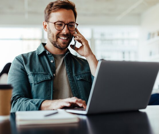 Mann am Laptop mit Smartphone am Ohr | © iStock/Jacob Lund Photography