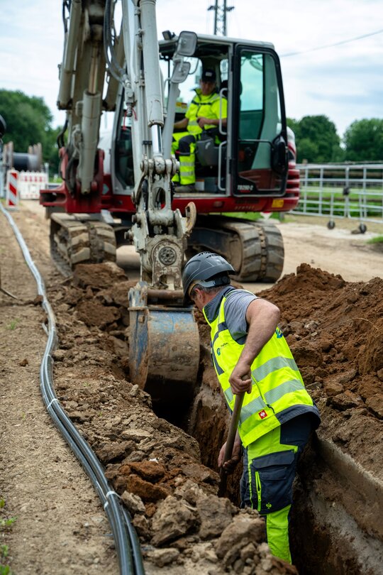 Bagger zieht einen Graben für das Erdkabel, ein Mitarbeiter steht mit einer Spate im Graben